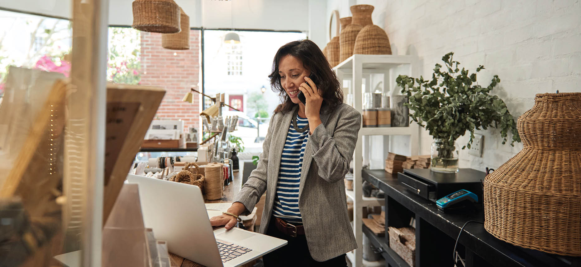 Smiling young Asian woman standing behind a counter in her stylish boutique working on a laptop and talking on a cellphone